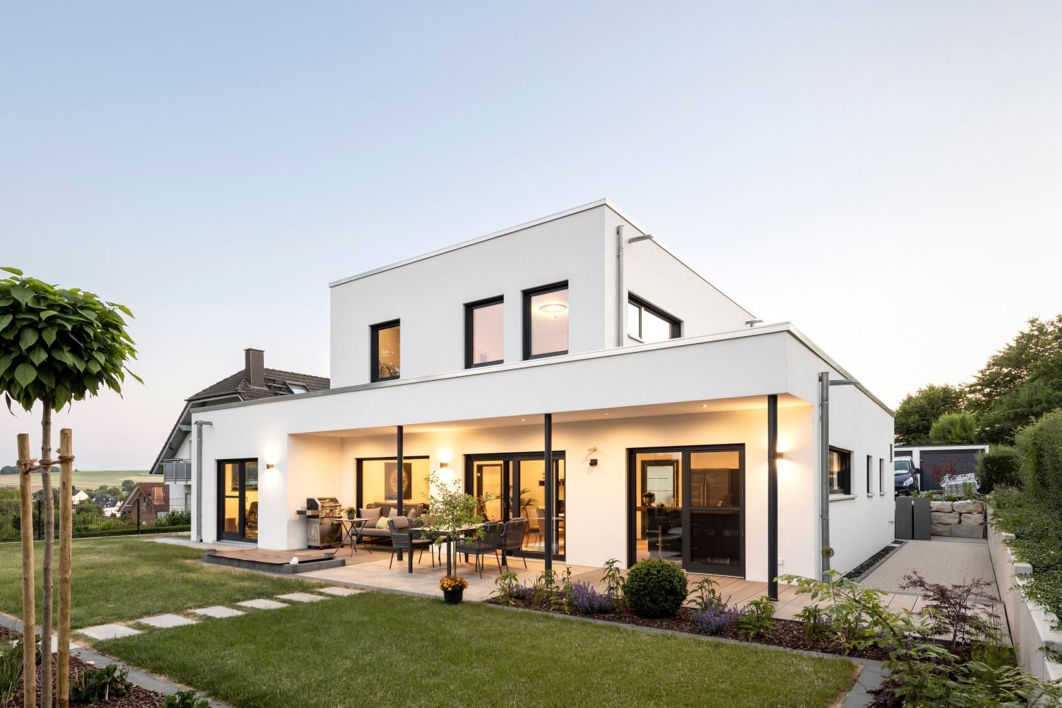Modernes frei geplantes FingerHaus Fertighaus mit klarer Architektur, Flachdach und großzügiger Terrasse bei Abendlicht, umgeben von einem gepflegten Garten.