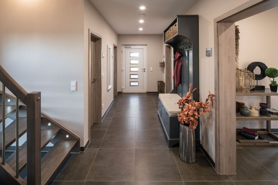 Geräumiger Flur mit dunklem Fliesenboden, Garderobe aus Holz mit Sitzbank und Ablagefächern im Musterhaus UNO in Leipzig. Im Hintergrund moderne Haustür mit Glaseinsätzen, links Holztreppe mit Beleuchtung und an der Wand dekorative Elemente.