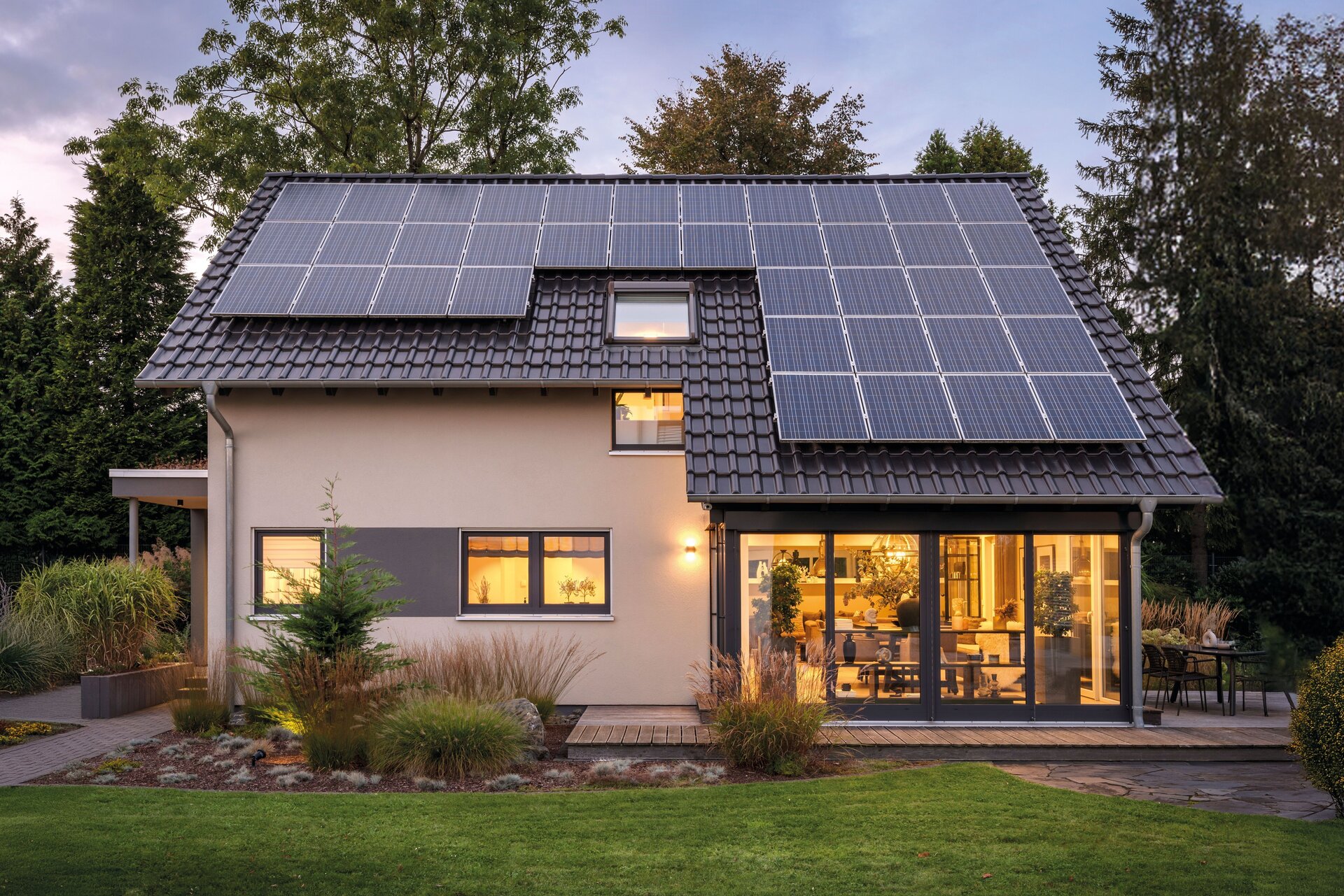 Musterhaus NEO von FingerHaus in Wuppertal bei Dämmerung, Einfamilienhaus mit Satteldach und Photovoltaikanlage, helle Putzfassade, große Glasfront mit Einblick in den beleuchteten Wohnbereich, gepflegte Gartenfläche und Bepflanzung im Vordergrund.
