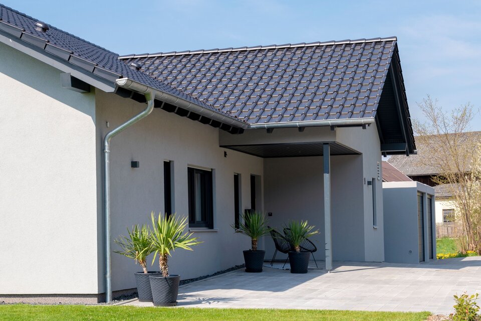 Seitenansicht eines FingerHaus NIVO 130 mit überdachtem Terrassenbereich, heller Fassade, dunklem Satteldach, Kübelpflanzen und gepflegtem Garten mit Rasenfläche und Zaun.