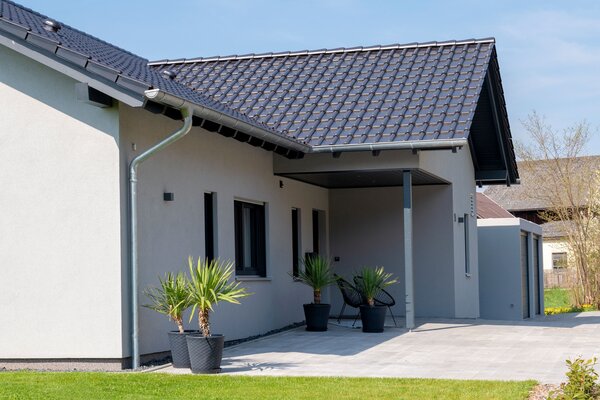 Seitenansicht eines FingerHaus NIVO 130 mit überdachtem Terrassenbereich, heller Fassade, dunklem Satteldach, Kübelpflanzen und gepflegtem Garten mit Rasenfläche und Zaun.