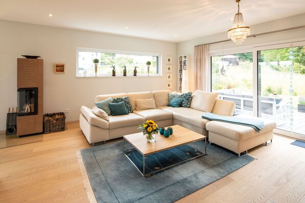 Gemütliches Wohnzimmer in einem frei geplanten FingerHaus mit heller Eckcouch, Kaminofen, Glastisch, großem Teppich und Zugang zur Terrasse durch bodentiefe Fenster.