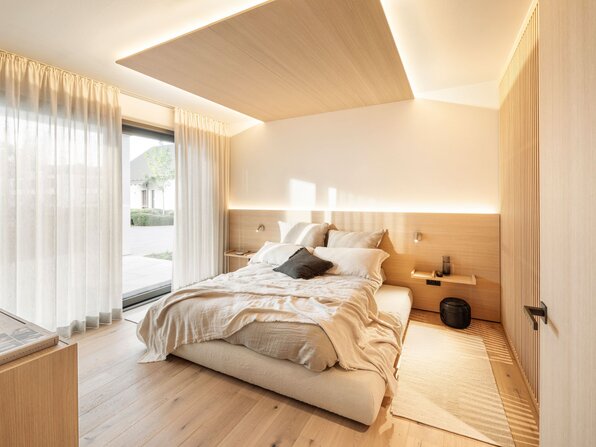 Schlafzimmer im FingerHaus Musterhaus NIVO in Frankenberg mit Doppelbett, Holzverkleidung, indirekter Beleuchtung und bodentiefem Fenster.