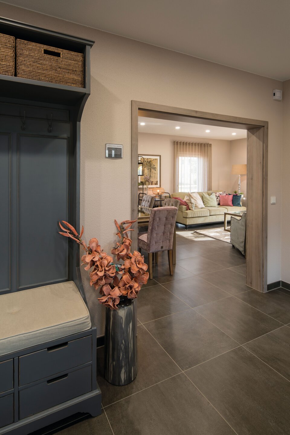 Flur mit dunklem Fliesenboden und grauer Garderobe, daneben Vase mit Trockenblumen. Durch die geöffnete Tür Blick ins Wohnzimmer mit Sofa, Tisch und großen Fenstern mit Vorhängen im Musterhaus UNO Leipzig.