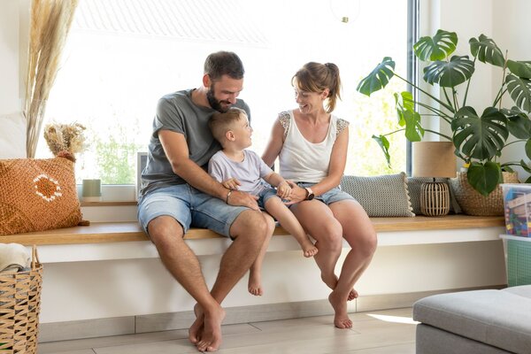 Familie sitzt entspannt auf einer breiten Fensterbank in einem hellen, freundlich gestalteten Wohnbereich mit Pflanzen und Kissen.