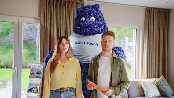 Ein Paar steht in einem Wohnzimmer, hinter ihnen das blaue Monster aus der FingerHaus-Kampagne „Das Unerwartete“ mit der Aufschrift „Bau-Pfusch“ – Symbol für die Angst vor schlechter Bauqualität.