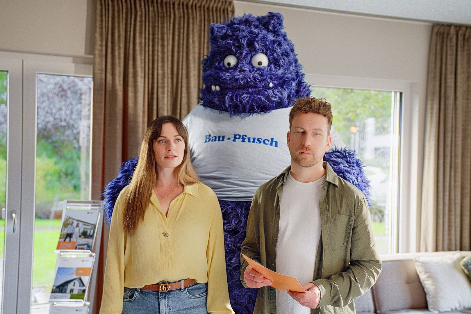 Ein Paar steht in einem Wohnzimmer, hinter ihnen das blaue Monster aus der FingerHaus-Kampagne „Das Unerwartete“ mit der Aufschrift „Bau-Pfusch“ – Symbol für die Angst vor schlechter Bauqualität.
