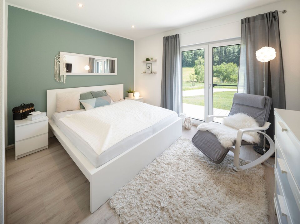 Gästezimmer mit Doppelbett, grün gestrichener Akzentwand und großem Fenster. Beistelltische mit Lampen, Sessel und Teppich schaffen eine gemütliche Atmosphäre.