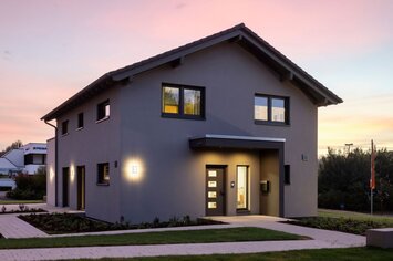 Musterhaus ARTIS 300 in Würzburg bei Abenddämmerung: Modernes zweigeschossiges Fertighaus mit klarer Architektur, beleuchtetem Eingangsbereich und gepflegtem Garten.