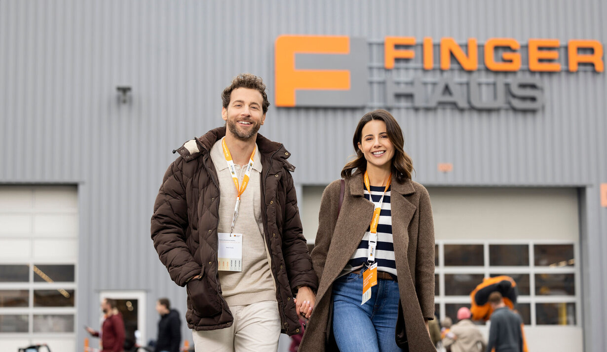 Ein Paar geht lächelnd über das Veranstaltungsgelände des FingerHaus-Bauinfotags in Frankenberg. Im Hintergrund ist eine Halle mit dem FingerHaus-Logo zu sehen, umgeben von weiteren Besuchern.