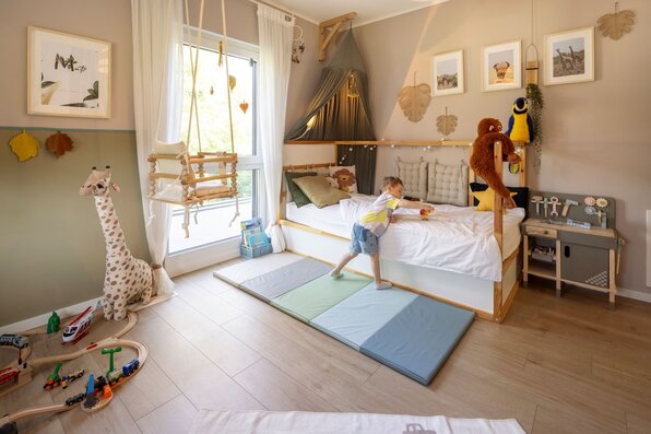 Hell und freundlich gestaltetes Kinderzimmer in einem frei geplanten FingerHaus mit großem Fenster, Holzfußboden und liebevollen Details. Ein Kind spielt fröhlich auf dem Bett, während Schaukel, Giraffenfigur und Spielzeugzüge Gemütlichkeit schaffen.