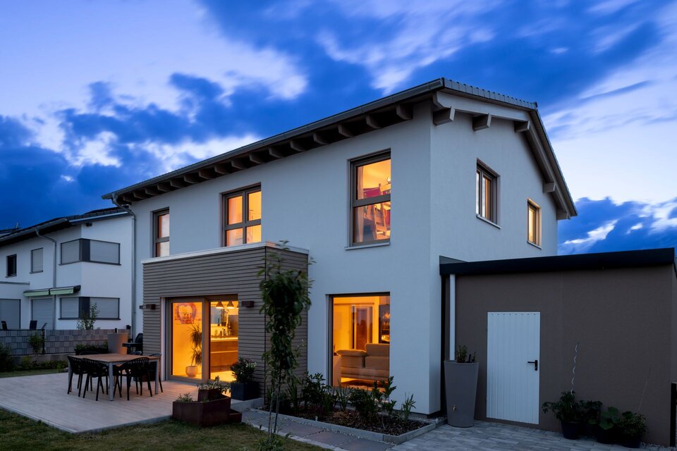 Zweigeschossiges weißes Einfamilienhaus in der Abenddämmerung mit beleuchteten Fenstern, Holzverkleidung und Terrasse.
