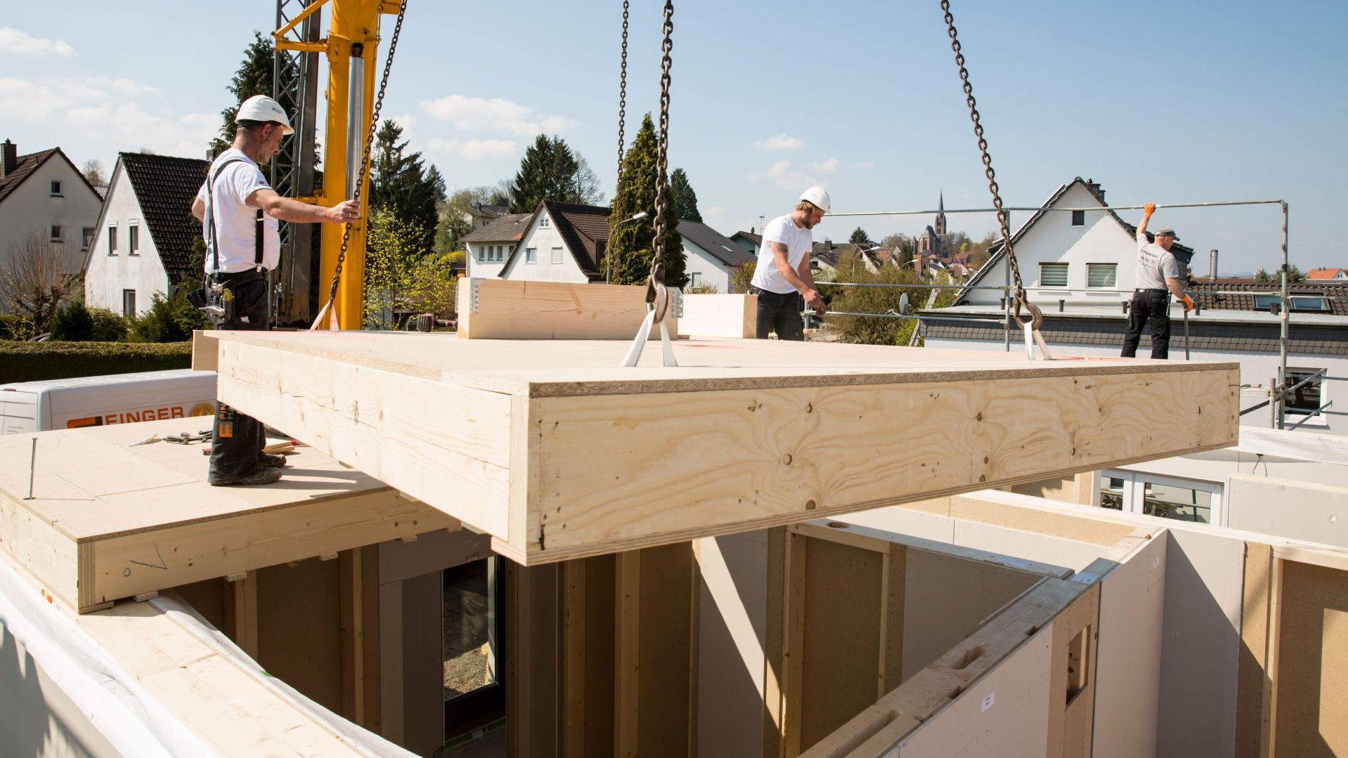 Mitarbeiter montieren ein Finger-Haus auf der Baustelle; ein großes Holzdeckenelement wird mit Ketten an einem Kran angehoben und von mehreren Arbeitern mit Schutzhelmen auf die vorbereiteten Wände gesetzt, im Hintergrund Wohnhäuser und blauer Himmel.