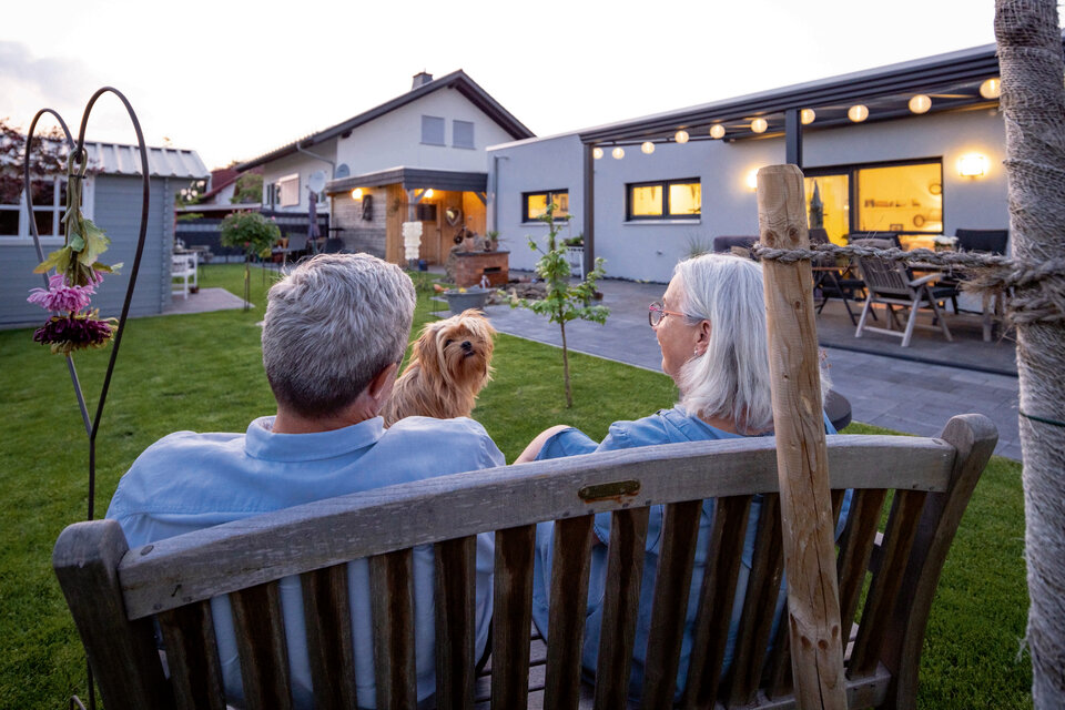Älteres Paar sitzt auf einer Gartenbank vor einem beleuchteten Bungalow mit Terrasse und Hund im Garten