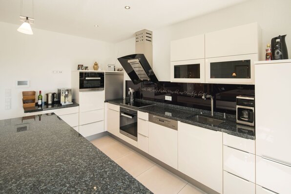 Moderne Einbauküche in einem frei geplanten FingerHaus mit weißen Fronten, dunkler Granit-Arbeitsplatte und hochwertigen Elektrogeräten.