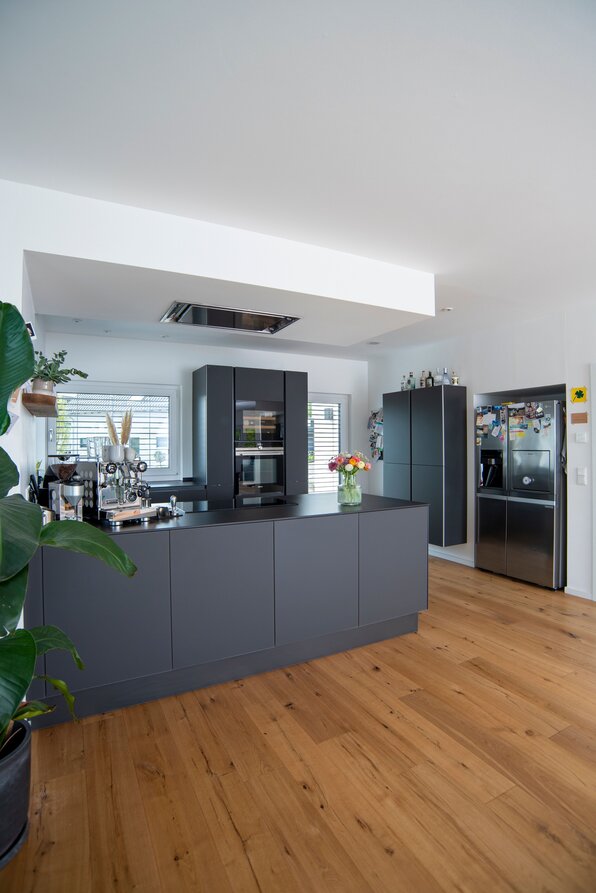 Moderne Küche in einem frei geplanten FingerHaus mit Kochinsel, dunklen Fronten und hellem Holzboden.