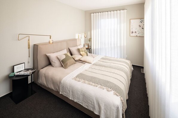 Schlafzimmer im Musterhaus TALO Fulda mit großem Bett, heller Bettwäsche, modernen Wandleuchten und lichtdurchfluteten Vorhängen.