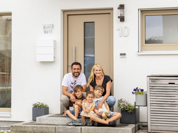 Eine fünfköpfige Familie mit zwei Erwachsenen und drei Kindern sitzt gemeinsam auf den Eingangsstufen vor einer Haustür.