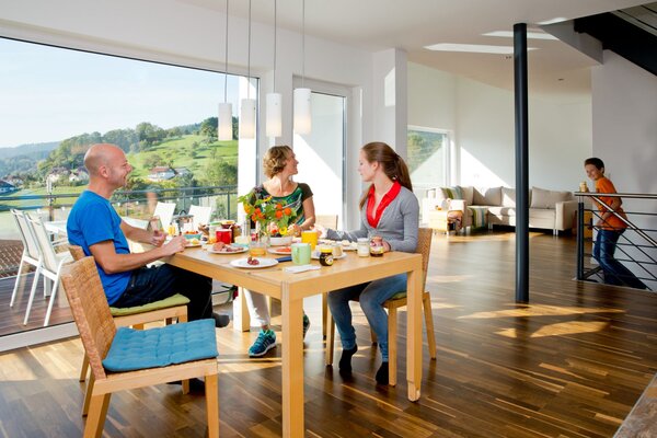 Familie sitzt beim Frühstück am Holztisch in einem hellen Essbereich mit Panoramafenstern und Blick auf grüne Hügel.