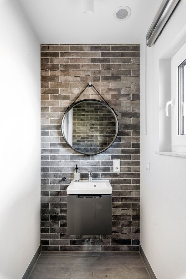 Modernes Gäste-WC im FingerHaus Musterhaus TALO in Babenhausen mit grauen Backsteinfliesen, kleinem Waschbecken und rundem Spiegel mit Lederriemen. Minimalistisches Design mit klaren Linien und hellen Akzenten.