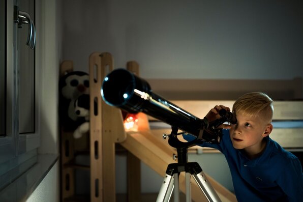 Junge schaut abends durch ein Teleskop aus dem Fenster seines Kinderzimmers.