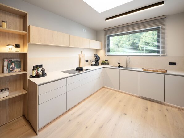 Küche im FingerHaus Musterhaus NIVO in Frankenberg mit L-förmiger Küchenzeile, weißen Fronten, Kochfeld, Spüle und großem Fenster.