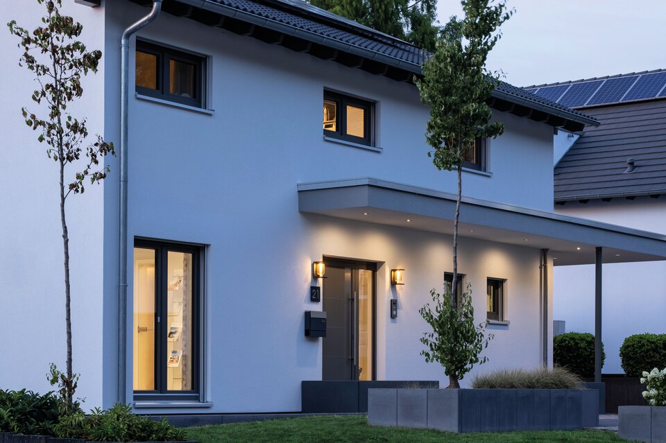 Abendansicht des FingerHaus Musterhauses SENTO 503 in Bad Vilbel mit beleuchtetem Eingangsbereich, moderner Fassade und überdachtem Eingang.