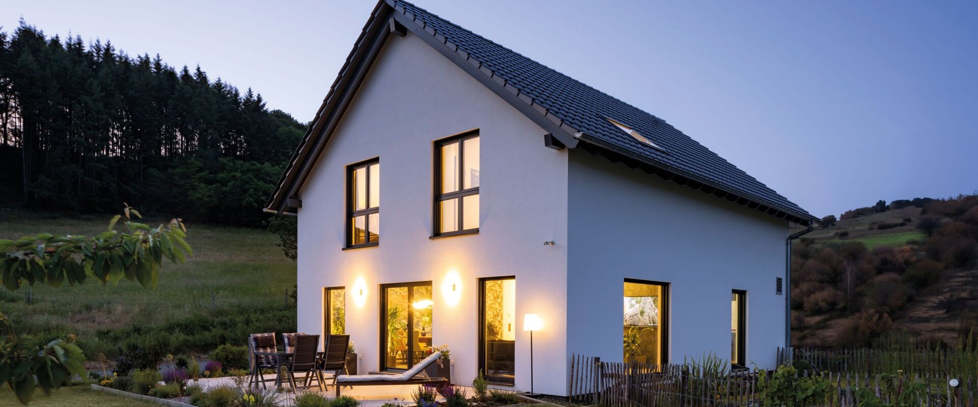 Zweigeschossiges Einfamilienhaus von FingerHaus in der Abenddämmerung mit beleuchteten Fenstern, Garten und Terrasse. Das Haus steht am Waldrand mit Blick auf eine hügelige Landschaft.
