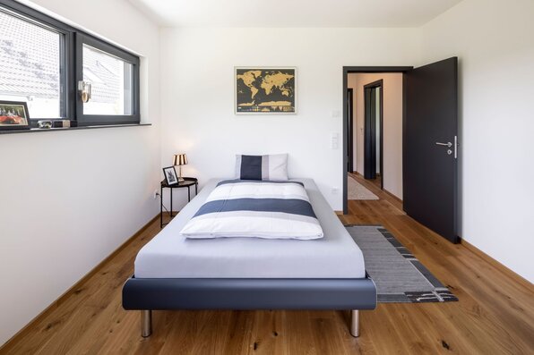 Gästezimmer in einem frei geplanten FingerHaus mit Einzelbett, Holzfußboden und schlichten weißen Wänden. Große Fenster sorgen für viel Tageslicht, dekoriert mit moderner Wandkarte und Nachttischlampe.