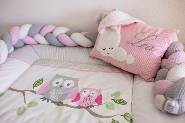 Detailaufnahme aus einem Kinderzimmer mit rosa Kissen, geflochtenem Bettschutz in Pastellfarben und Bettwäsche mit Eulenmotiv auf einem Ast.