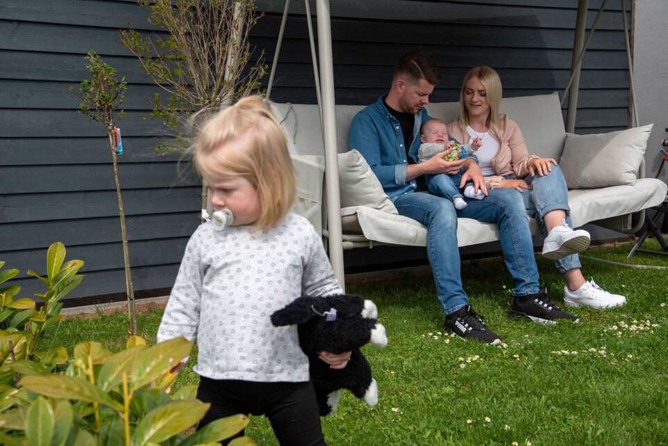 Eine Familie sitzt auf einer Gartenschaukel vor ihrem FingerHaus, während ein kleines Kind mit einem Stofftier im Vordergrund auf dem Rasen spielt.