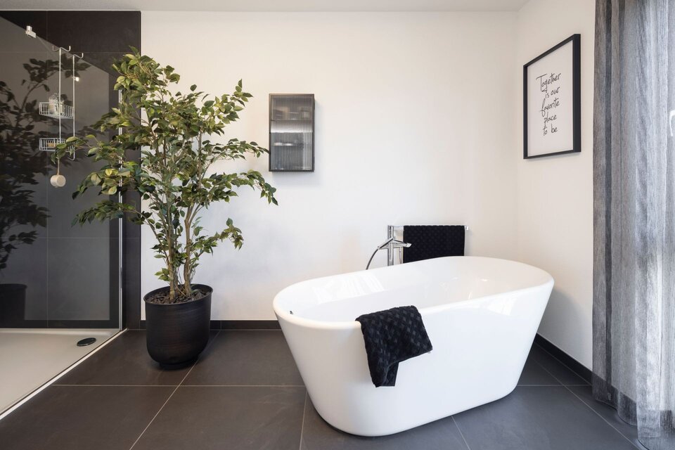 Modernes Badezimmer mit freistehender weißer Badewanne auf dunklem Fliesenboden im Musterhaus TALO Fulda. Daneben schwarze Armatur, Handtuch, große grüne Pflanze im Topf und Glasduschwand im Hintergrund.