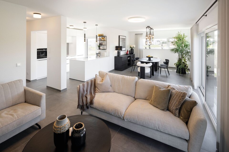 Offener Wohnbereich im Musterhaus TALO in Fulda mit gemütlicher Sofalandschaft, moderner Küche und Essplatz in hellen Farben.