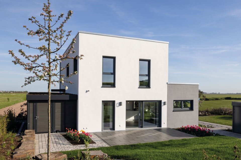 Außenansicht des FingerHaus-Musterhauses TALO in Fulda mit kubischer Architektur, Flachdach, weißer Fassade, großen bodentiefen Fenstern, Terrasse, Rasenfläche, Gartenbeeten, Baum und freistehendem Geräteschuppen unter blauem Himmel.