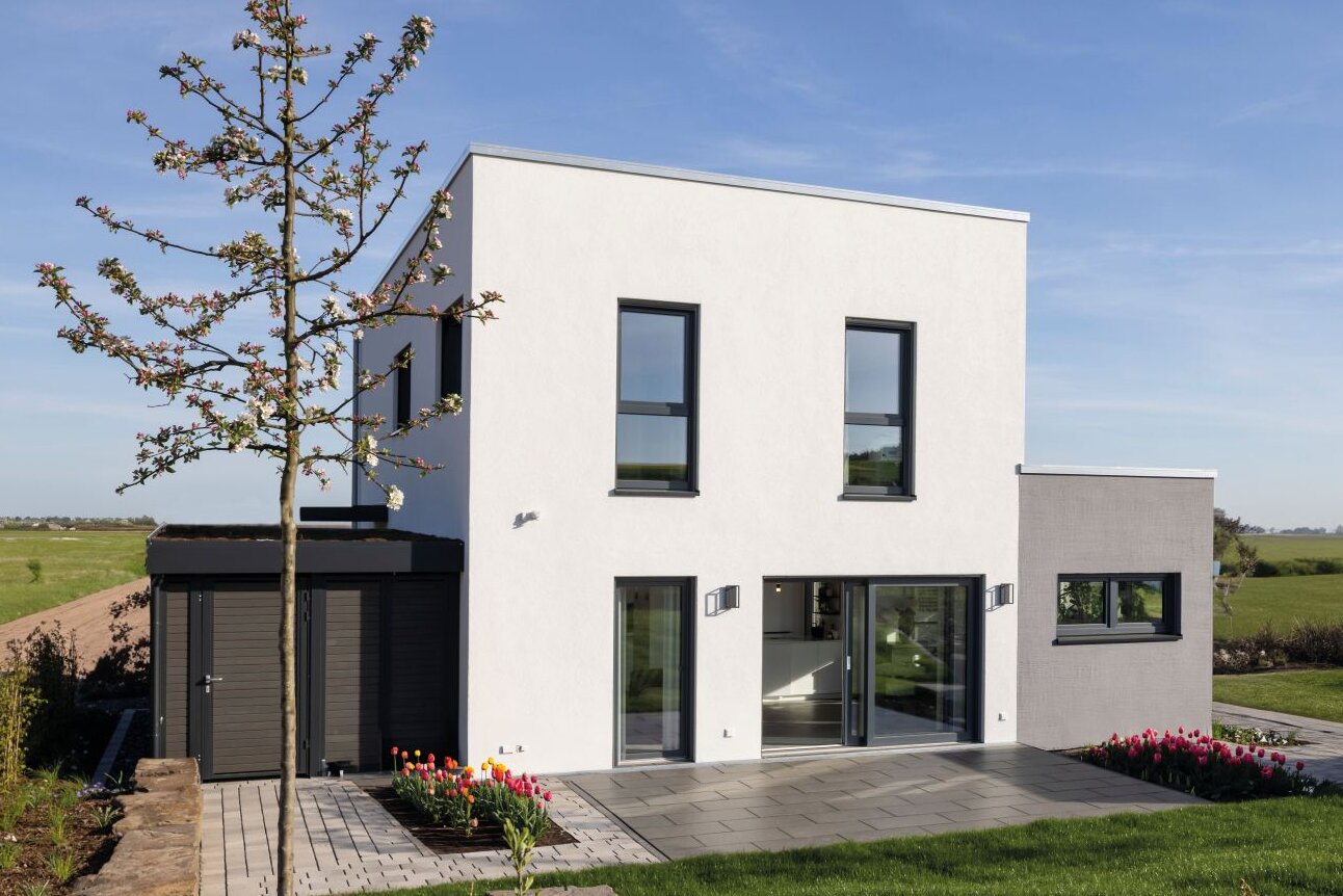 Außenansicht des FingerHaus-Musterhauses TALO in Fulda mit kubischer Architektur, Flachdach, weißer Fassade, großen bodentiefen Fenstern, Terrasse, Rasenfläche, Gartenbeeten, Baum und freistehendem Geräteschuppen unter blauem Himmel.