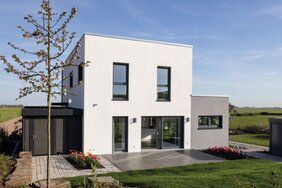Außenansicht des FingerHaus-Musterhauses TALO in Fulda mit kubischer Architektur, Flachdach, weißer Fassade, großen bodentiefen Fenstern, Terrasse, Rasenfläche, Gartenbeeten, Baum und freistehendem Geräteschuppen unter blauem Himmel.