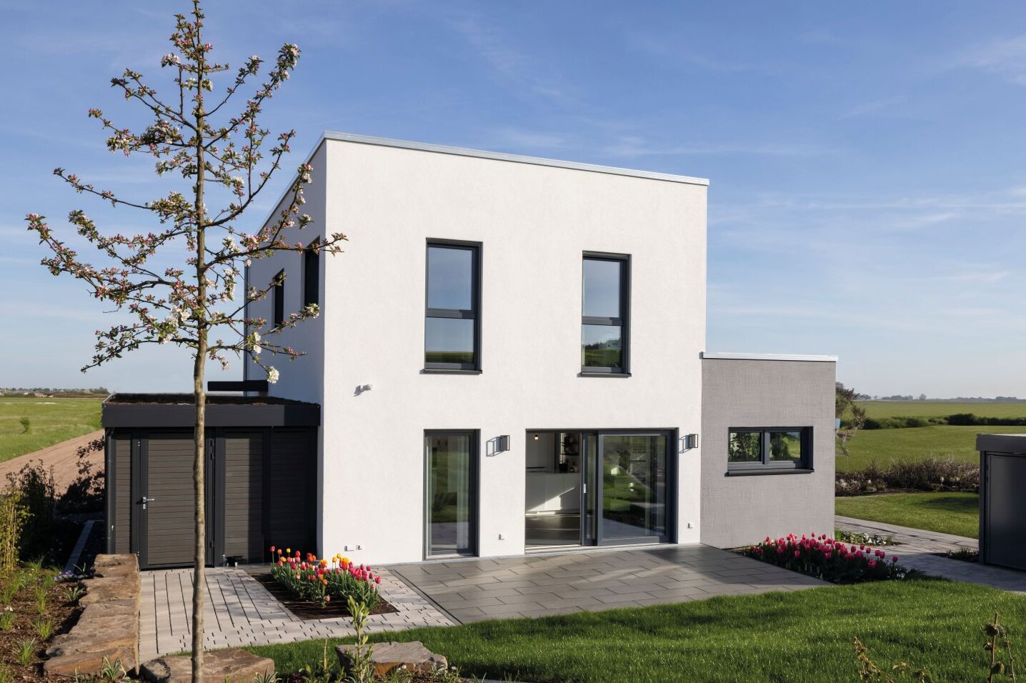 Außenansicht des FingerHaus-Musterhauses TALO in Fulda mit kubischer Architektur, Flachdach, weißer Fassade, großen bodentiefen Fenstern, Terrasse, Rasenfläche, Gartenbeeten, Baum und freistehendem Geräteschuppen unter blauem Himmel.