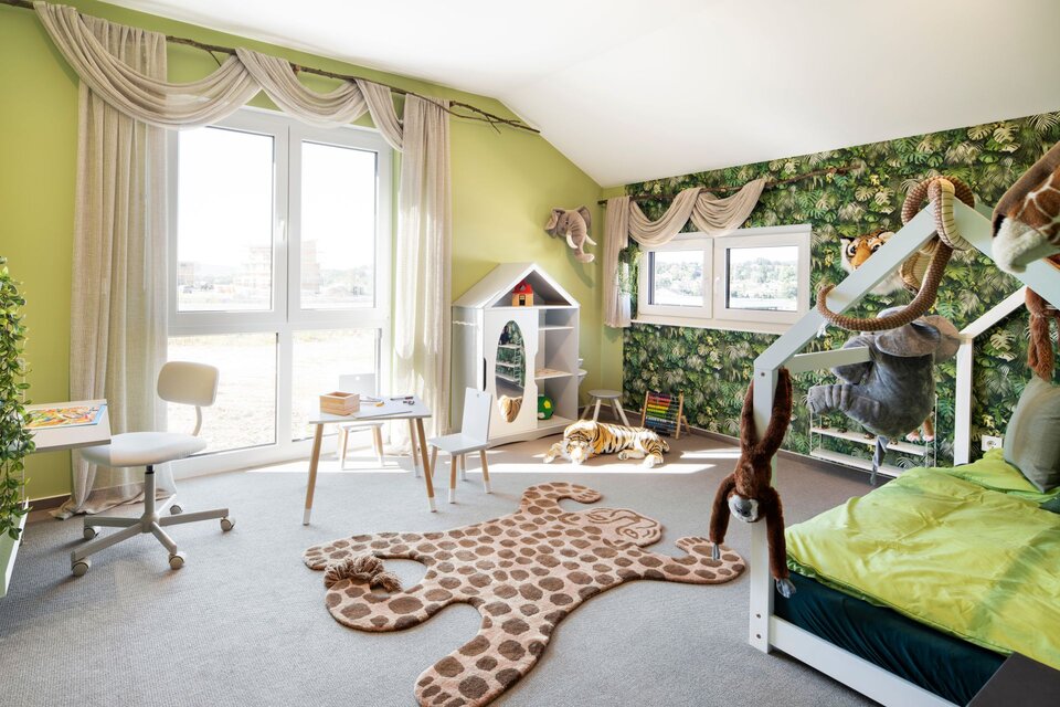 Kinderzimmer mit Dschungelmotiv, grün gestrichenen Wänden und Tapete mit Blättern. Hausbett, Spielmöbel, großes Fenster und Teppich in Tierform schaffen eine verspielte Atmosphäre.