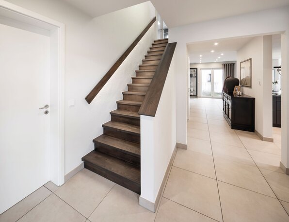 Moderner heller Flur mit dunkler Holztreppe, weißen Wänden und hellen Bodenfliesen, rechts ein schwarzer Schrank und ein Bild an der Wand