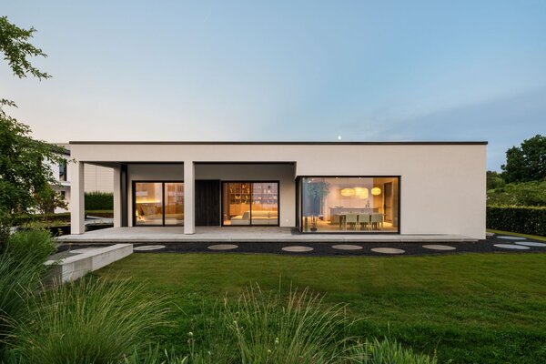 Rückansicht des Musterhauses NIVO in Frankenberg mit moderner Bungalowarchitektur, überdachter Terrasse, großzügigen Fensterfronten und beleuchteten Wohnräumen in der Abenddämmerung.