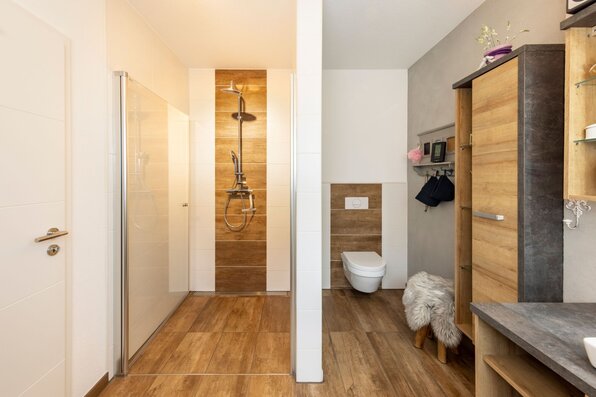 Modernes Badezimmer mit großer begehbarer Dusche, Holzbodenfliesen, hellem Waschtisch und WC im Hintergrund.
