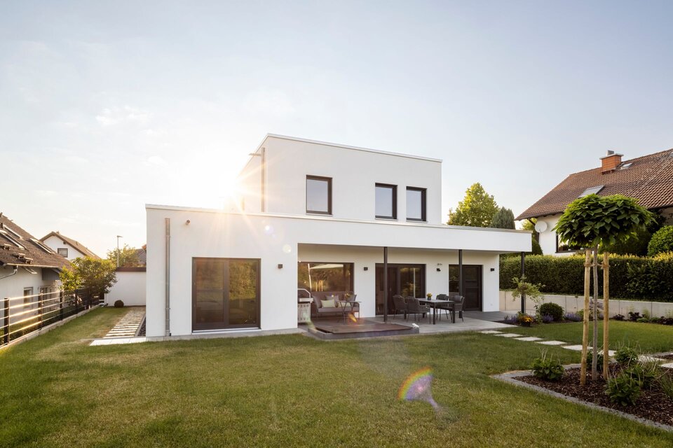 Moderne Außenansicht eines frei geplanten FingerHaus im Bauhaus-Stil mit weißer Fassade und klaren Linien. Große Fenster und eine überdachte Terrasse mit Sitzbereich öffnen sich zum gepflegten Garten.