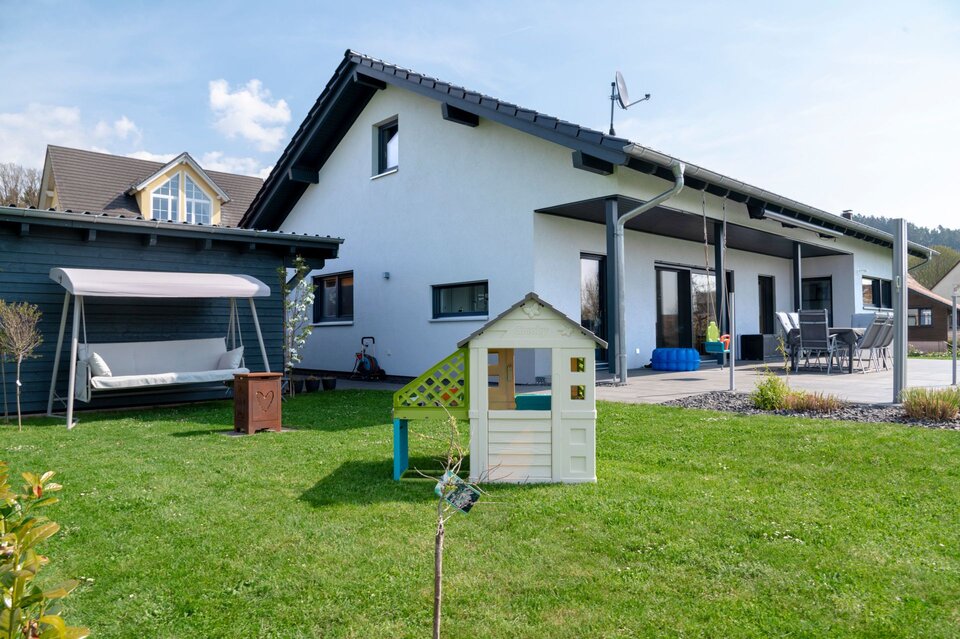 Rückansicht eines FingerHaus NIVO 130 mit überdachter Terrasse, großer Rasenfläche, Kinderspielhaus und Gartenschaukel im Hofbereich.