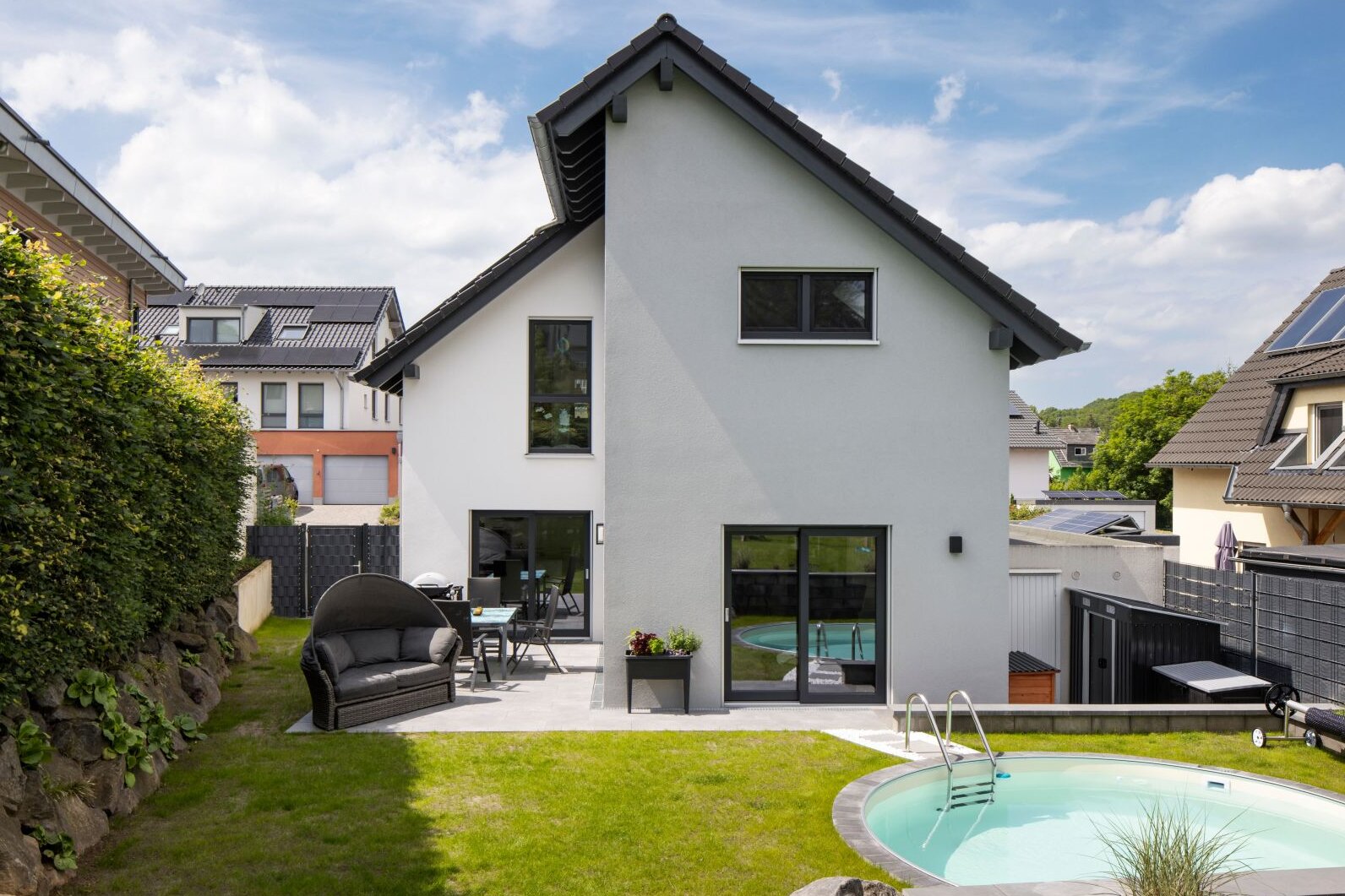 Rückansicht eines modernen Einfamilienhauses mit weißer Fassade, dunklem Satteldach und gepflegtem Garten. Im Vordergrund befinden sich ein runder Pool, Rasenfläche und Gartenmöbel auf einer Terrasse.