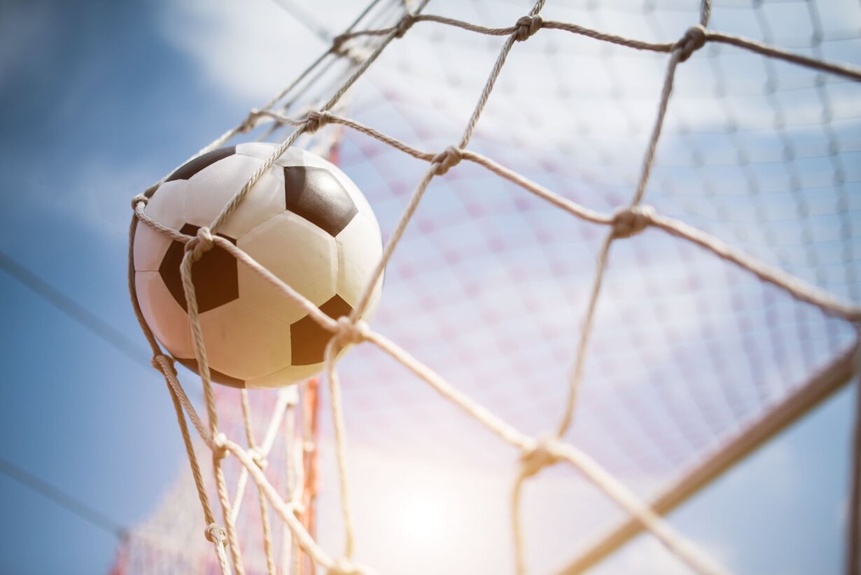 Fußball, der im Netz eines Tores hängt, vor blauem Himmel mit leichtem Sonnenlicht