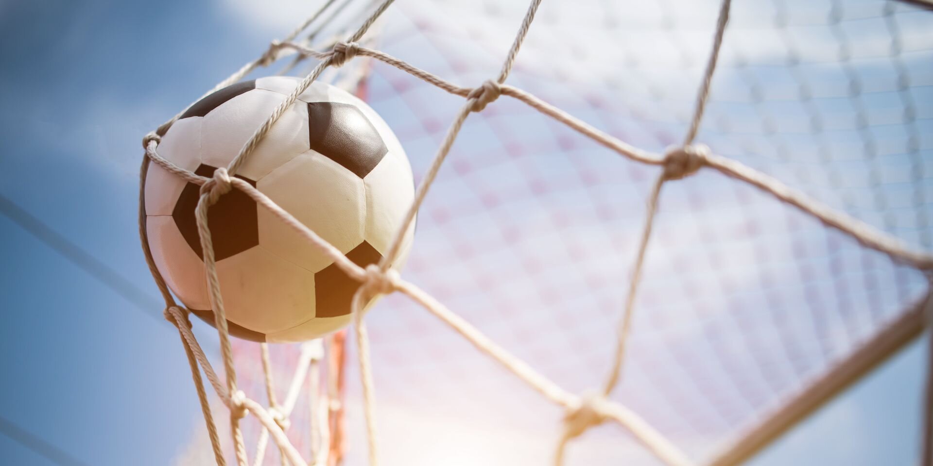 Fußball, der im Netz eines Tores hängt, vor blauem Himmel mit leichtem Sonnenlicht