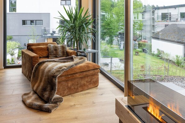 Gemütliche Leseecke in einem modernen FingerHaus-Haus mit großem Sessel, Felldecke, Gaskamin und bodentiefen Fenstern mit Blick ins Grüne.