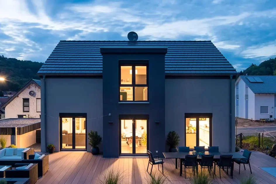Modernes Fertighaus mit dunklem Putz, großen Fenstern und beleuchteter Terrasse am Abend