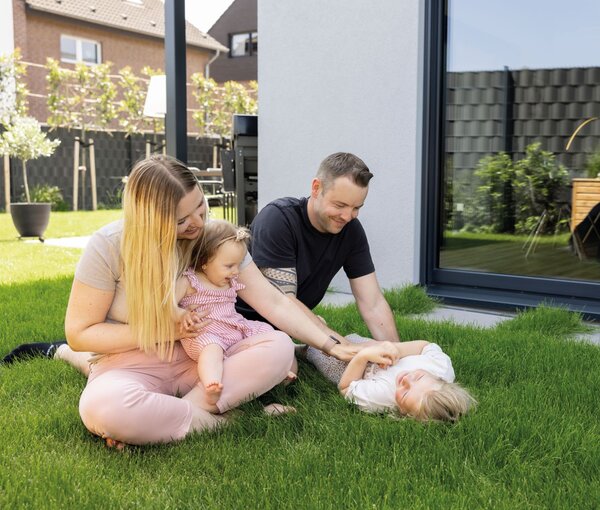 Familie mit zwei Kindern sitzt und spielt auf dem Rasen im Garten vor einer großen Fensterfront