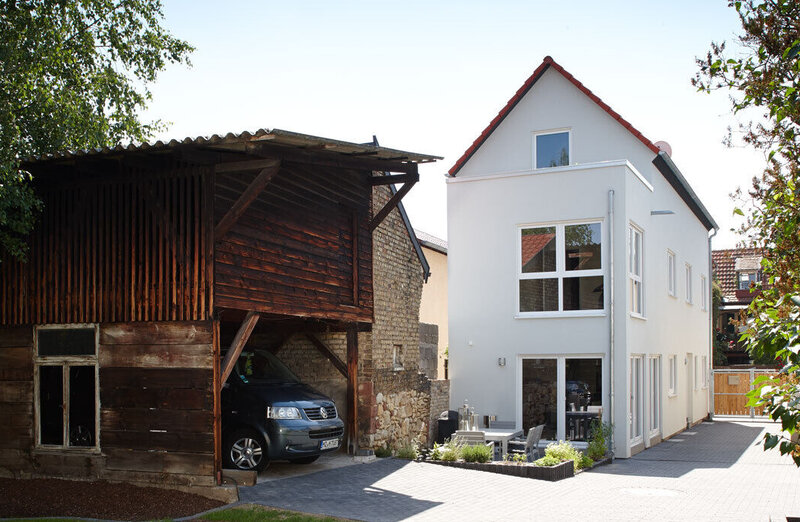 Ein modernes, zweigeschossiges Haus mit großen Fenstern neben einem traditionellen Holzgebäude. Ein Auto steht in der Einfahrt, umgeben von grünem Rasen und Bäumen.